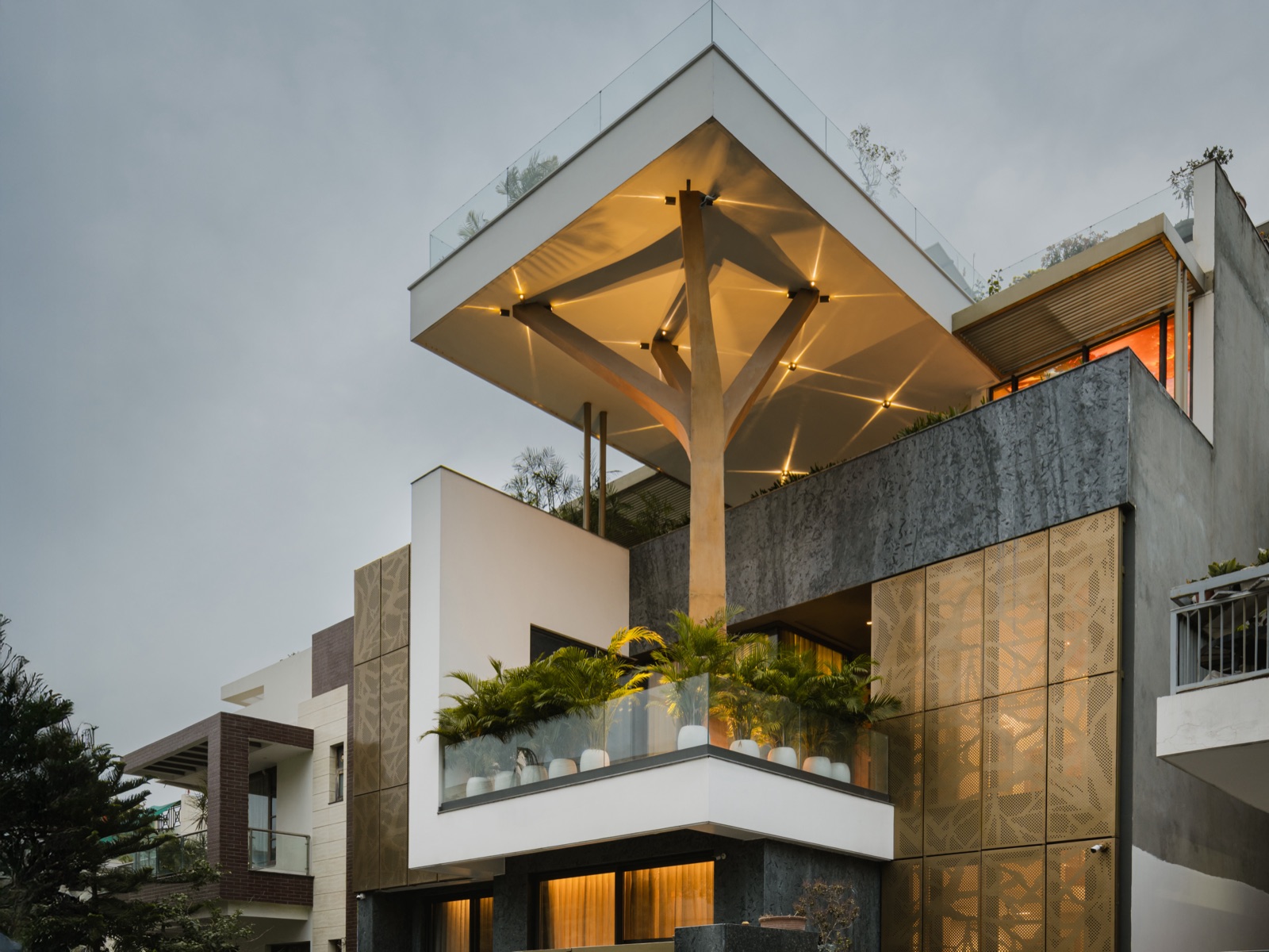 Photograph of a contemporary house at dusk: a white cantilevered roof canopy carried on a branching Y-shaped column, with small warm downlights in the canopy soffit casting star-shaped glints, a planted balcony with a glass railing, perforated gold metal facade panels and warm light behind curtained ground-floor windows