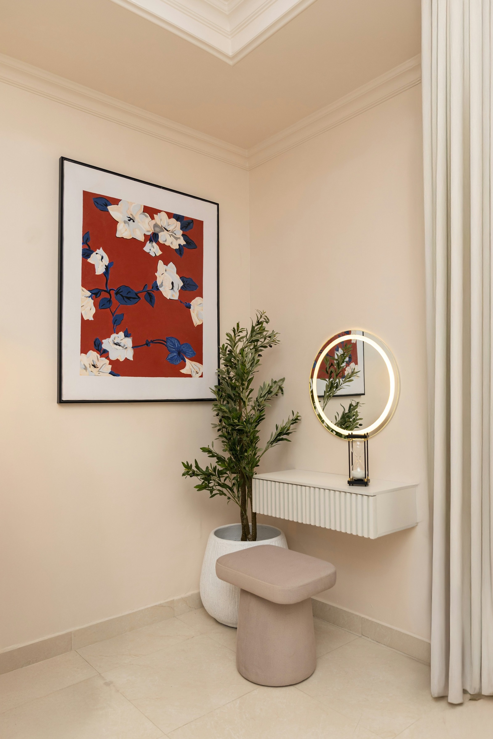 A corner of a cream bedroom with a slim wall-hung white dressing shelf with a fluted front, a round mirror with a glowing LED ring above it, a soft pink stool below, a potted olive tree and a large red and blue floral print on the wall.