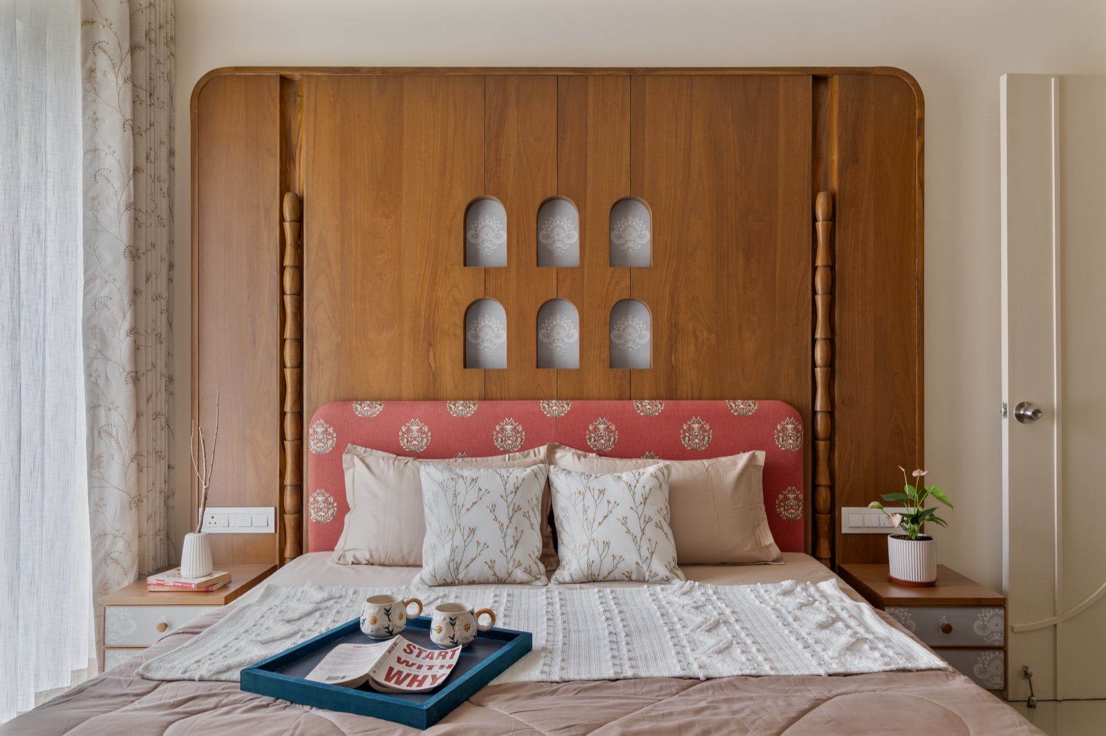 A bedroom with a double bed set against a full-height wooden wall panel with six small arched niches, a coral fabric headboard with a block-print motif, matching bedside tables with drawers, sheer curtains on the left and a white door on the right.