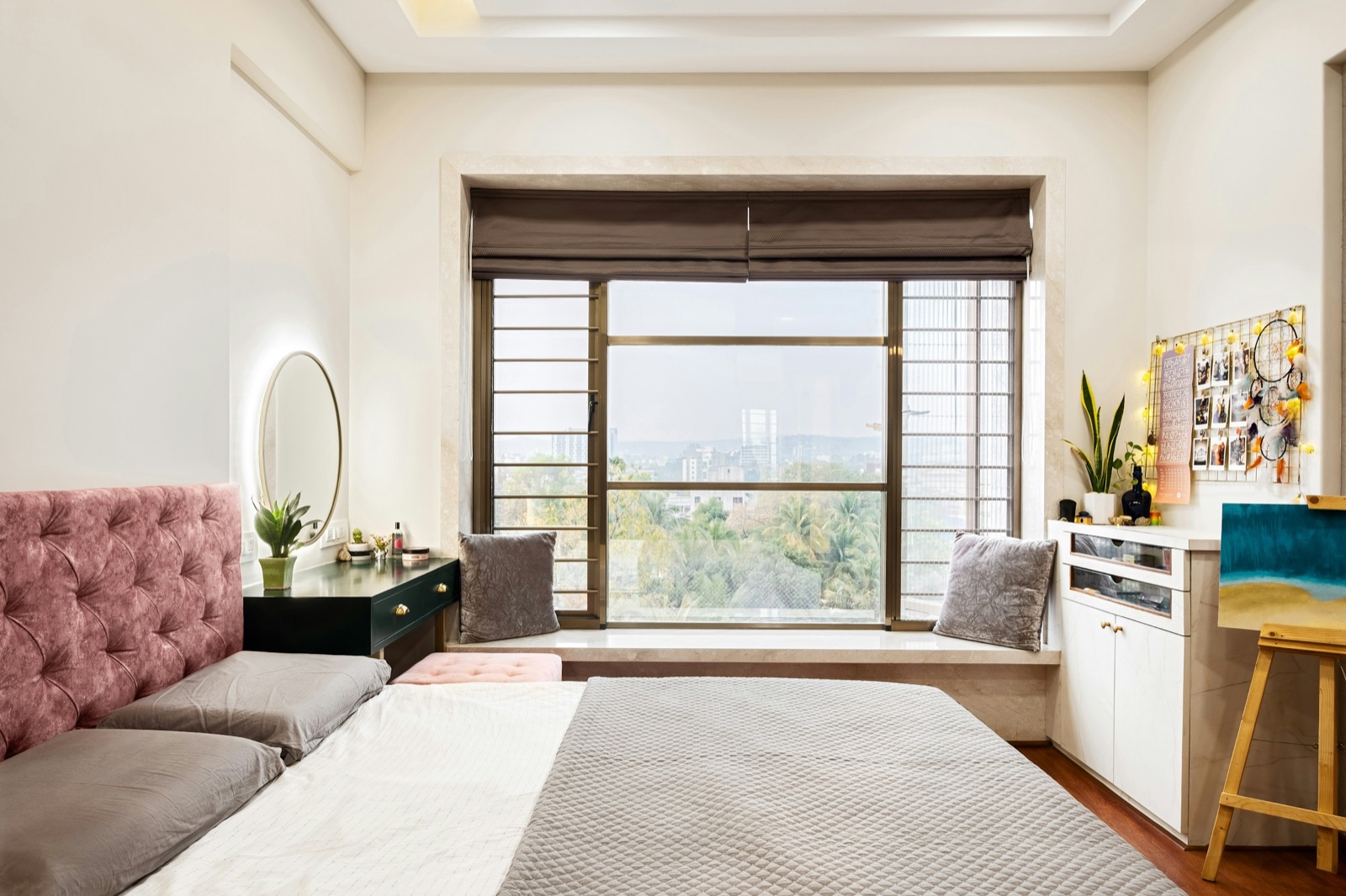 A bright bedroom in a Pune flat: a pink tufted headboard and grey quilted bed in the foreground, a dark green dressing table with a round backlit mirror beside the bed, a stone window seat with two cushions under a large window with a brown roman blind and a view over trees and buildings, and a white cabinet with an easel on the right.