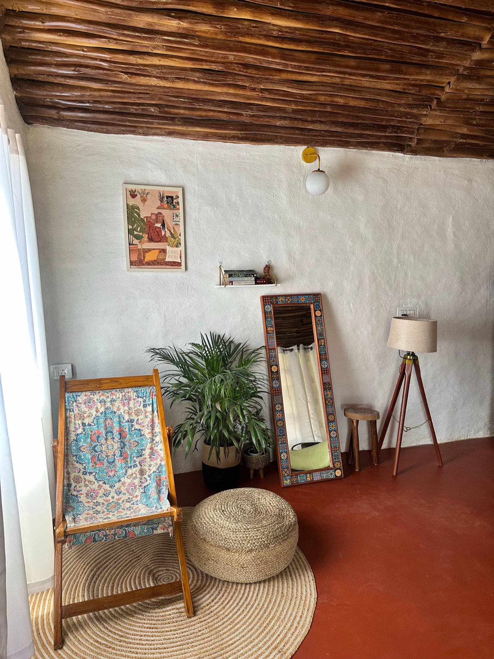 A corner of a whitewashed room in Kanthalloor, Kerala, with a timber-pole ceiling and a red floor: a wooden folding chair with a printed cotton sling, a round jute rug and jute pouf, an areca palm in a basket, a tile-framed mirror leaning on the wall, a tripod floor lamp, a small wooden stool, a globe wall light and a slim wall shelf with books