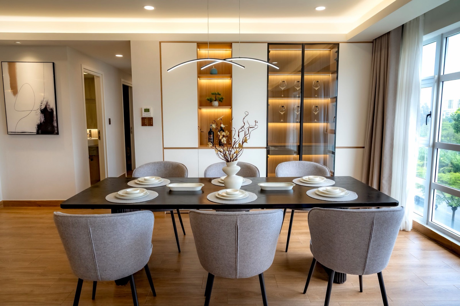 Dining room with a full-height white built-in unit: a tall glass-door showcase with warm LED light along each shelf displaying wine glasses, beside a lit open niche of walnut shelves, behind a dark dining table set for six with grey upholstered chairs