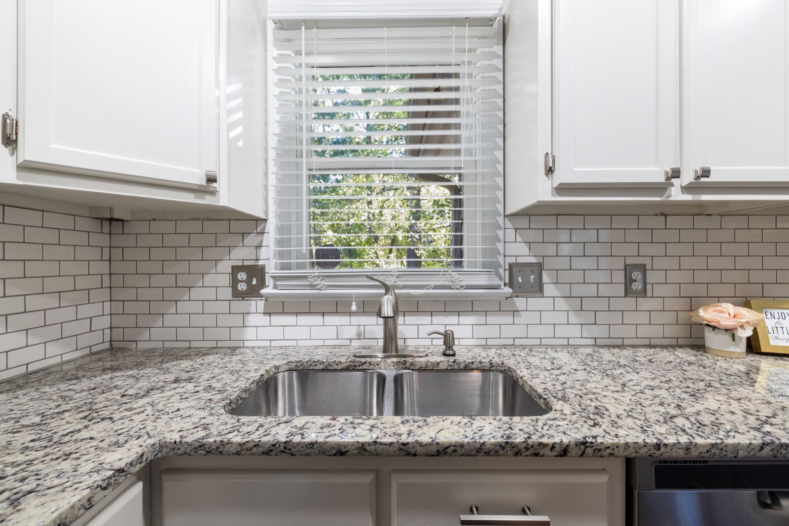 White glossy subway tiles with dark grey grout lines laid in brick bond from a speckled grey granite counter up to the underside of white wall cabinets, around a window with white blinds above a double steel sink.