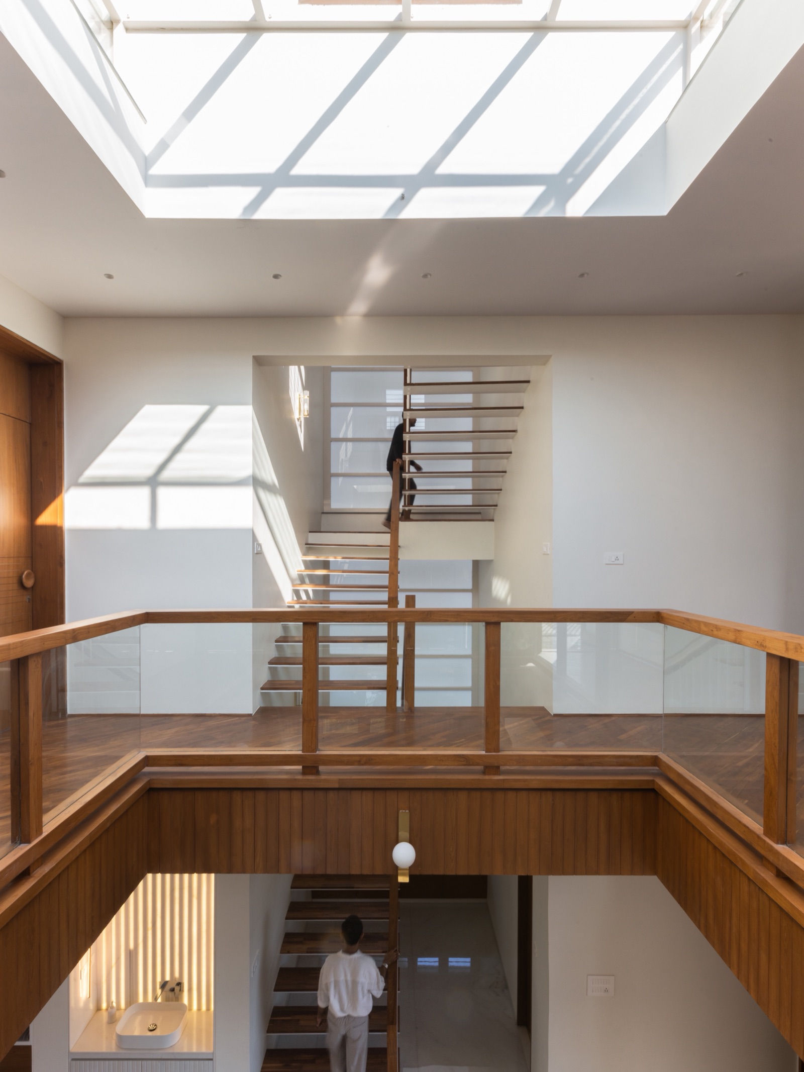 View across a double-height void with a timber and glass railing towards a dog-leg staircase of thin timber treads with open risers, lit by a large skylight, in an Ahmedabad bungalow