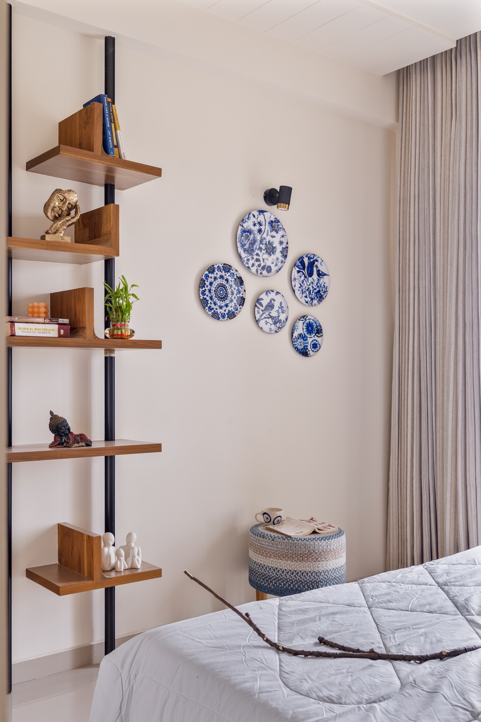 Walnut-finish wall shelves of different depths, some with an upright bookend side, mounted on two black vertical poles in a cream-walled bedroom in Vadodara, holding a few books, a brass face sculpture, a small plant, a reclining Buddha figurine and white figurines, beside a cluster of blue-and-white decorative plates on the wall