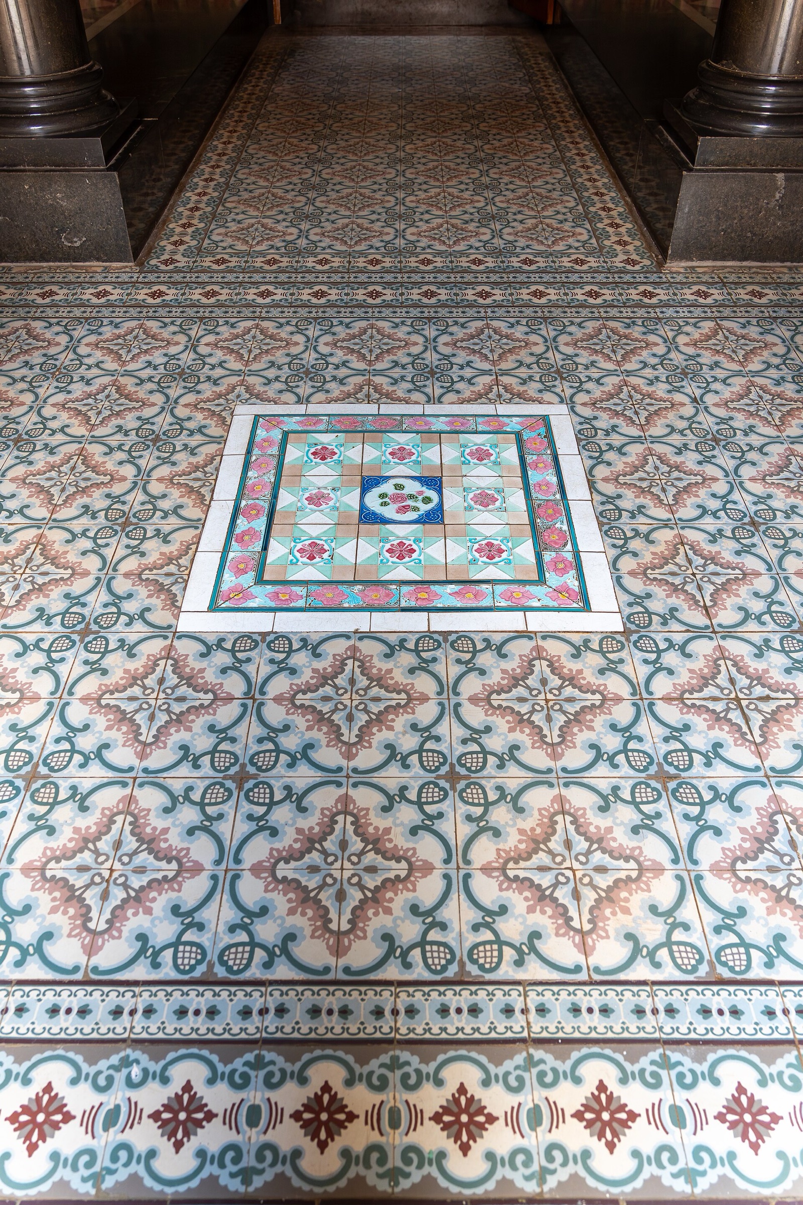 A floor of square cement tiles in pale blue, green and rose floral patterns, laid as a long runner between dark polished pillars, with a bordered central panel of glazed floral tiles and a patterned border band in the foreground.