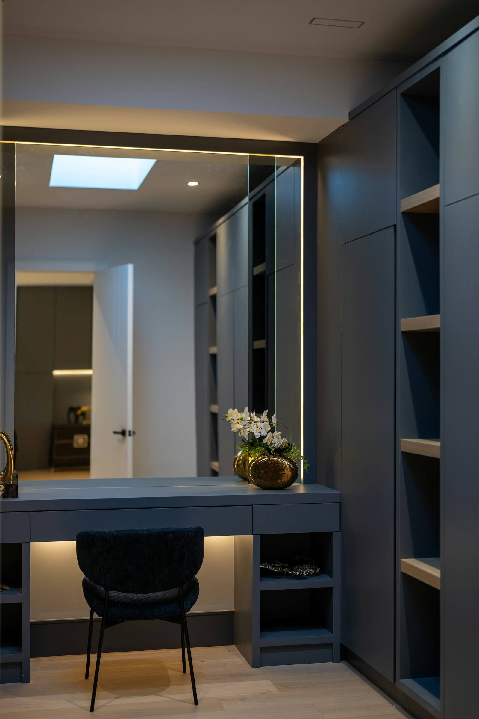 A built-in dressing table in matt slate blue joined to a run of tall wardrobe units and open shelves in the same finish, with a wall-wide mirror edged by a thin strip of light, a shallow drawer under the counter, open cubbies beside the knee space, a dark upholstered chair and a gold vase of white orchids.