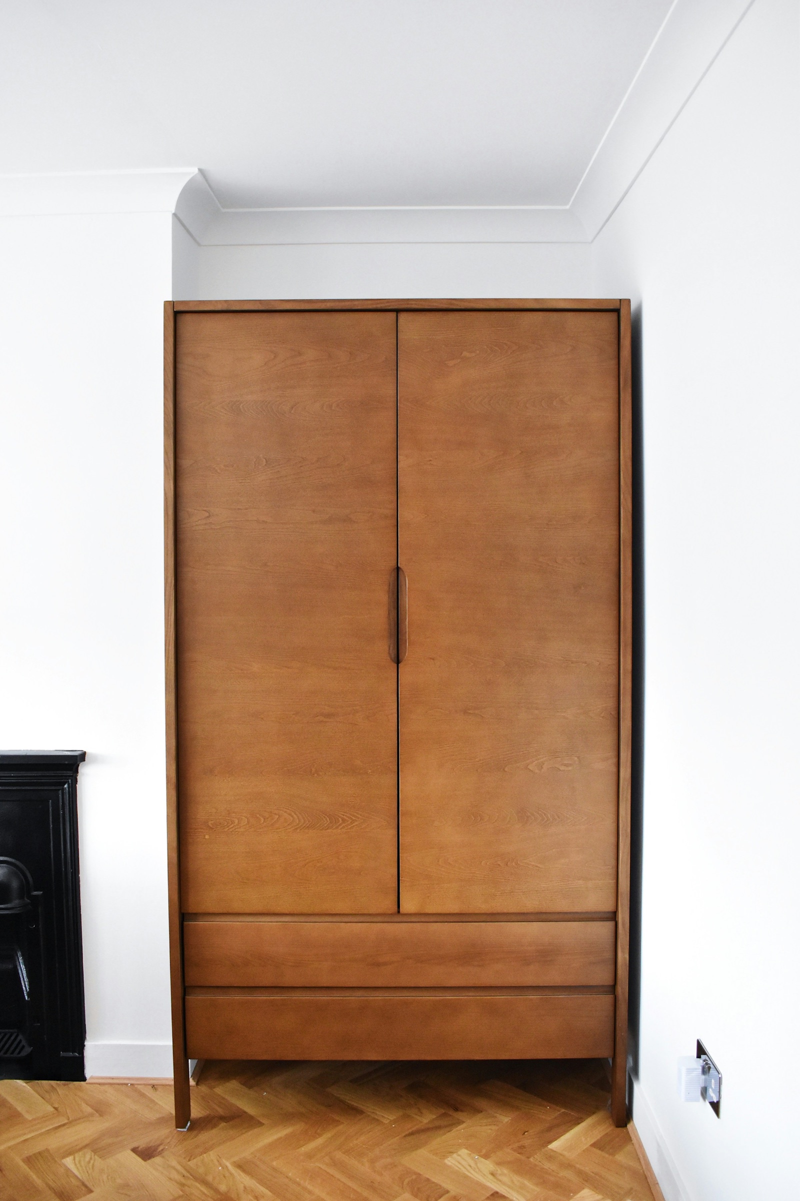 Freestanding two-door wardrobe in mid-brown wood with two drawers below, standing on legs in a white-walled alcove beside a black fireplace surround, with herringbone parquet flooring and empty wall above it up to the cornice