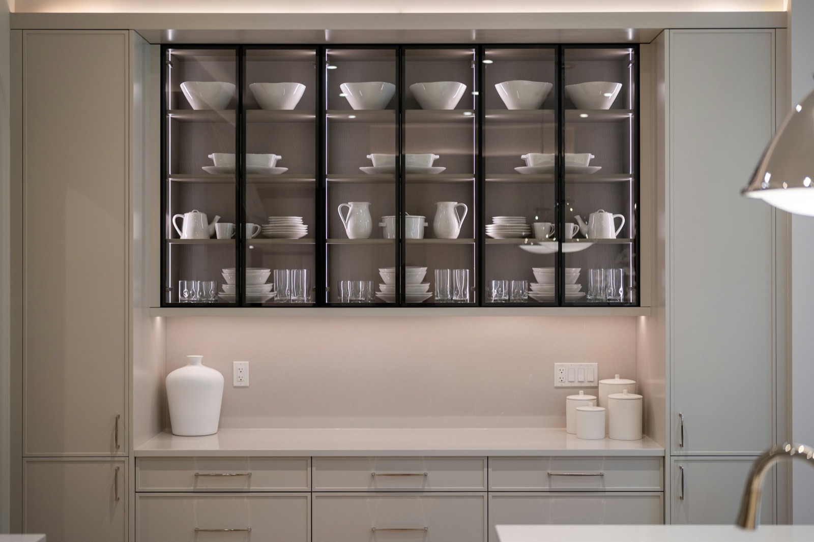 A built-in crockery unit along a wall: a row of six black-framed clear glass doors with lit glass shelves holding white bowls, serving dishes, jugs, teapots, stacked plates and tumblers, set above a pale stone counter with white canisters and a vase, with pale grey drawers below and tall grey cabinets on both sides.