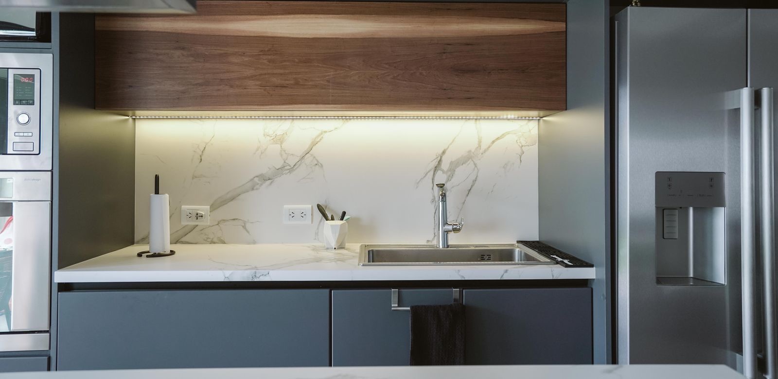 A sink run with a marble-look backsplash lit by an LED strip under a walnut-finish wall unit, between a built-in oven and a steel refrigerator