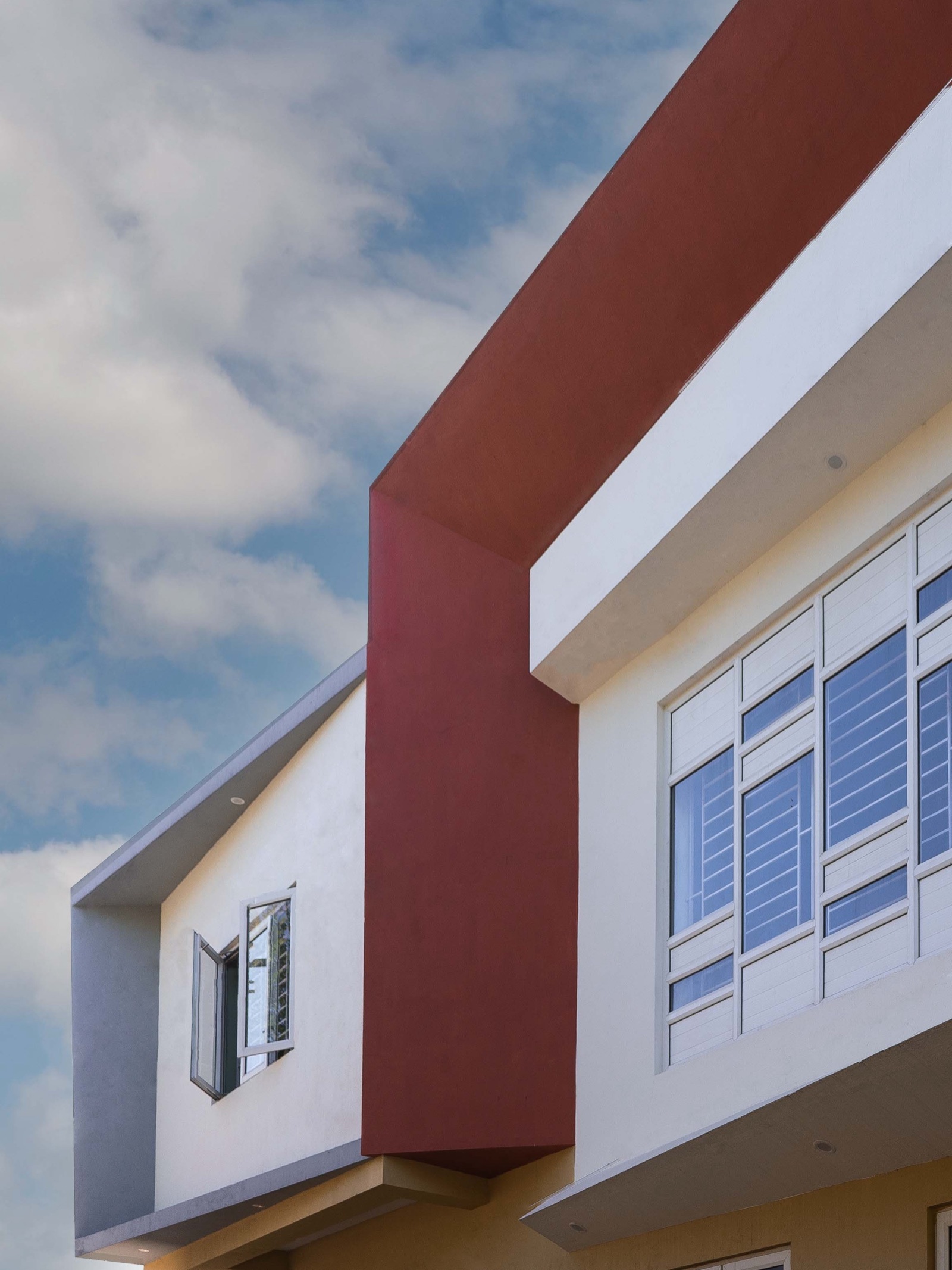 Upper floors of a house seen from below against a blue sky: a white volume with a slanted grey frame, a deep terracotta-red fin that folds over the roof edge, and a white box framing a large grid of windows