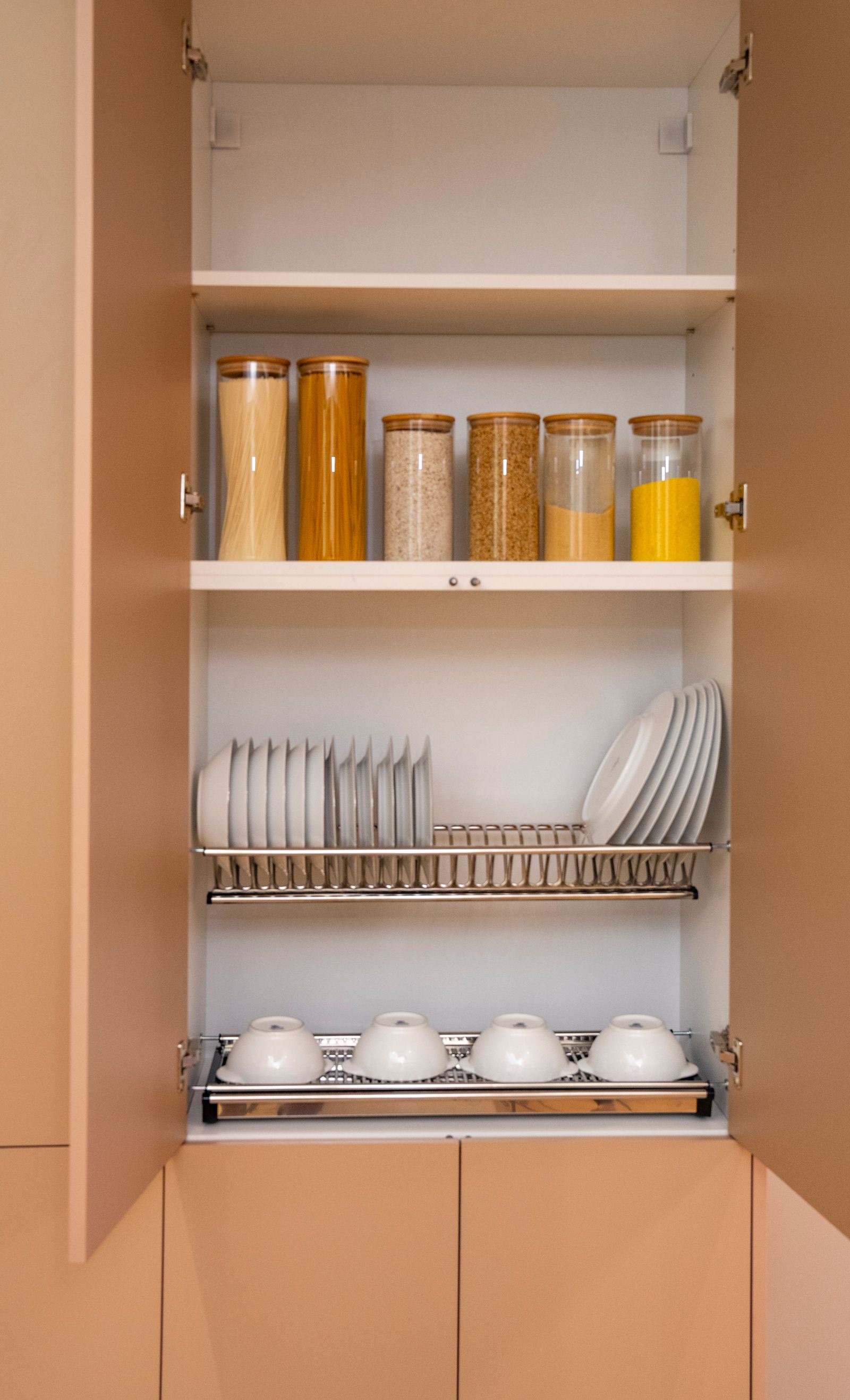 An open cabinet with storage jars on a shelf, a stainless steel plate rack holding white plates upright, and a bowl rack with a drip tray below