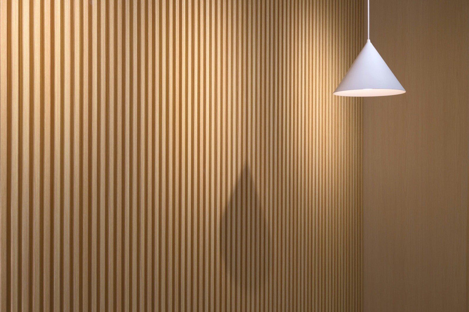 Close photograph of a wall of narrow light-oak-finish fluted strips with deep shadow lines between them, meeting a flat panel in the same finish, with a white cone pendant lamp hanging in front.