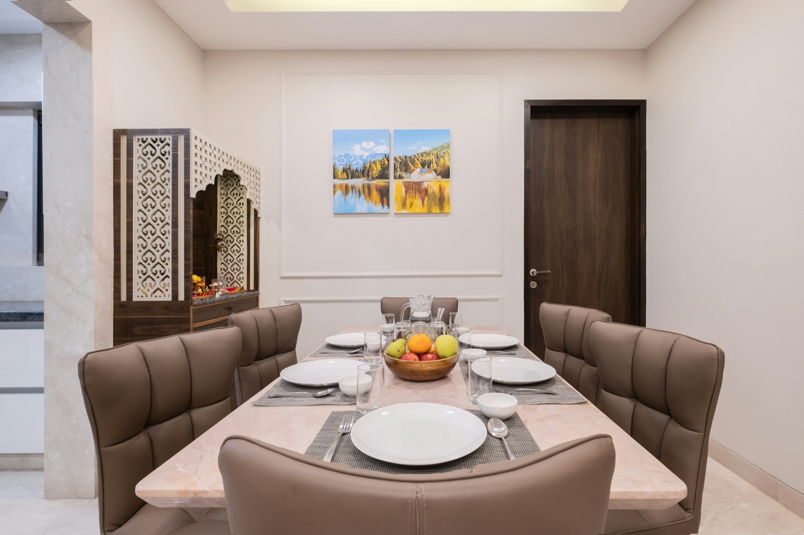 A dining table for six with a pale stone top and brown upholstered chairs, set with plates, glasses and a bowl of fruit. To its left stands a wooden puja unit with white jaali shutters and an arched jaali frame, lit inside, beside the kitchen; a panelled wall with a two-part landscape painting and a dark wooden bedroom door are behind.