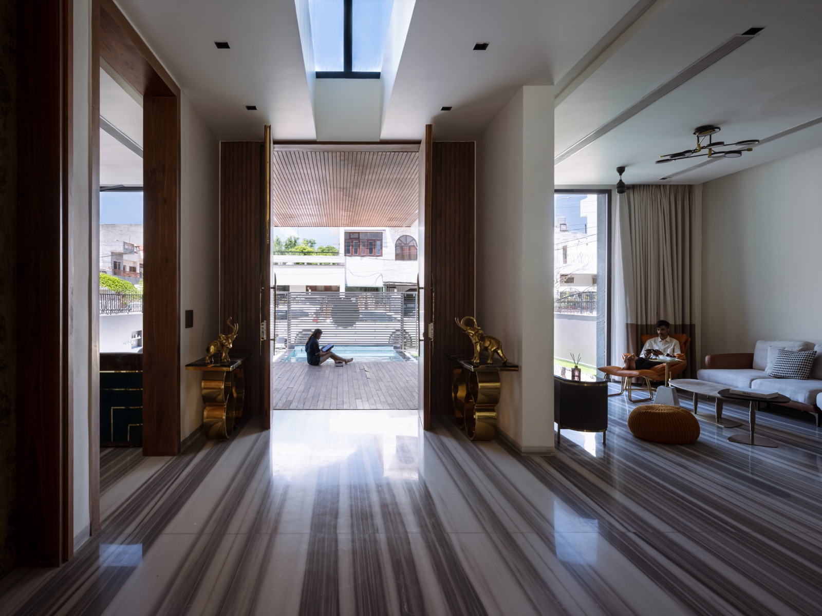 A double-height foyer with striped marble flooring, where two tall dark wooden door leaves stand open to a timber deck and pool, flanked by gold elephant figures on brass consoles under a skylight