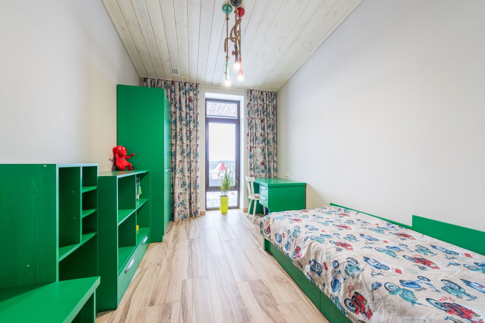 A long, narrow kids room with all its furniture in one green: a single bed with drawers below along the right wall, a small desk and white chair by a full-height glass door, a tall wardrobe and two open shelf units with drawers along the left wall, on a light wood-look floor.