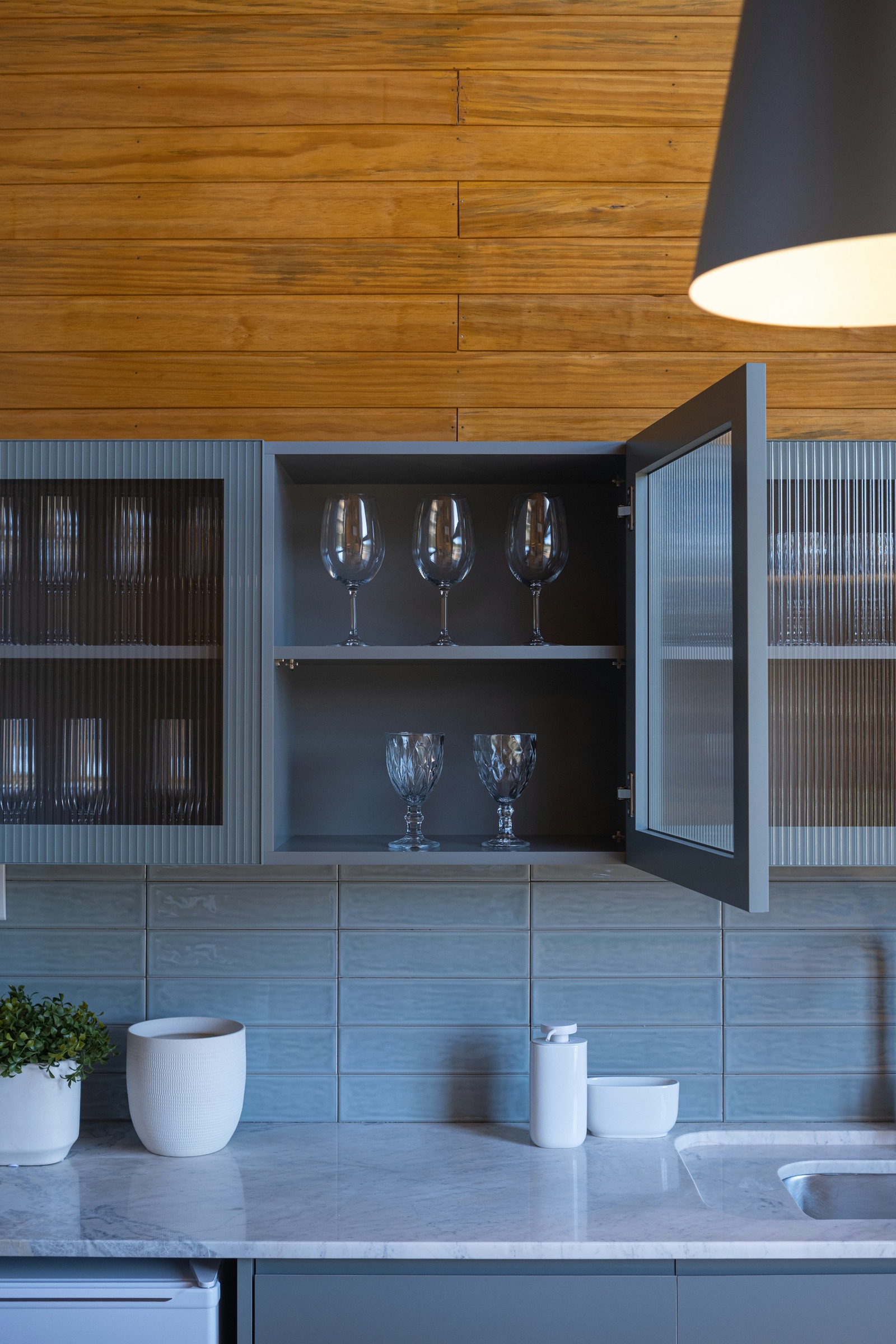 Grey kitchen wall cabinets with ribbed reeded glass doors above a marble counter and a blue tiled backsplash. One door stands open to show stemmed wine glasses on two shelves, while the closed reeded doors on either side blur the glassware behind them into vertical stripes.