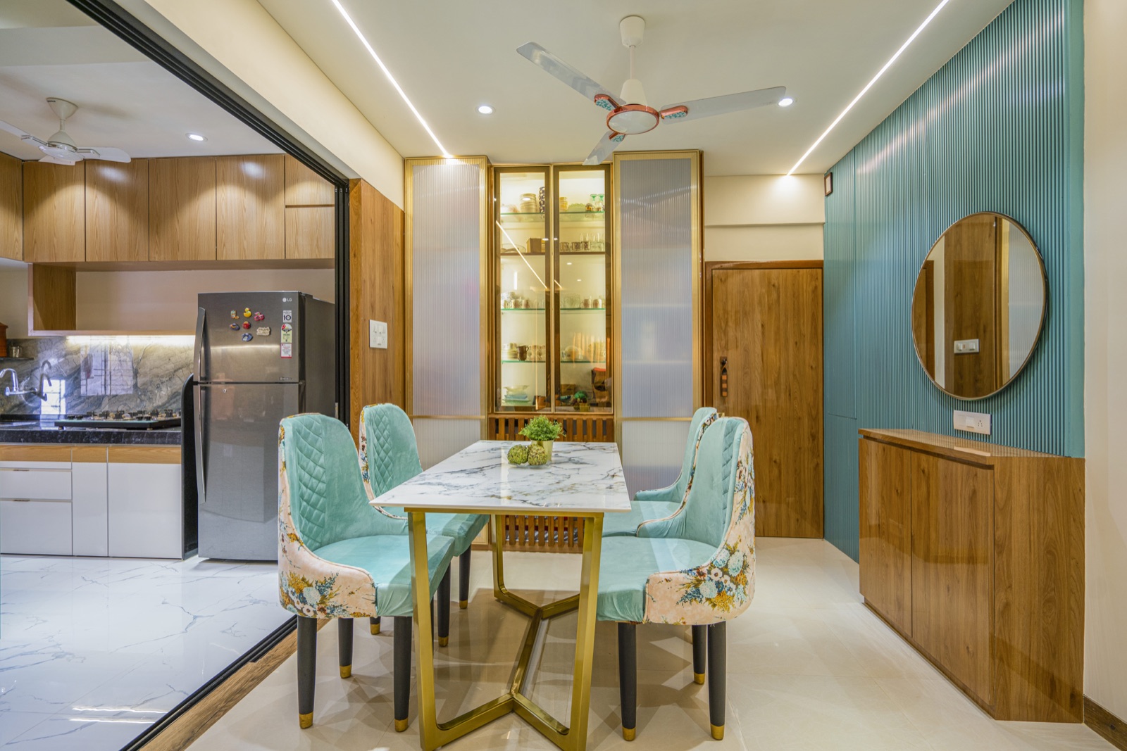 Dining area in a compact Mumbai apartment: a marble-topped table with four mint upholstered chairs, a tall glass-fronted display unit flanked by fluted glass shutters, a slim wooden cabinet below a round mirror, and an open kitchen with wood-finish wall units behind a black-framed glass partition