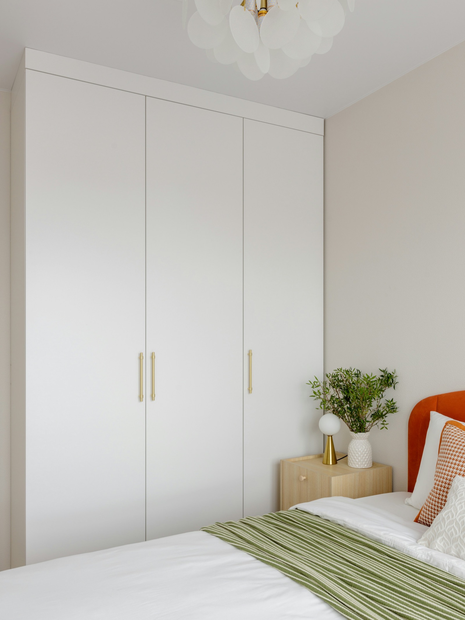 White flat-panel built-in wardrobe with three full-height doors and brass bar handles, beside a bed with an orange headboard and a light wood bedside table in a small bedroom with beige walls