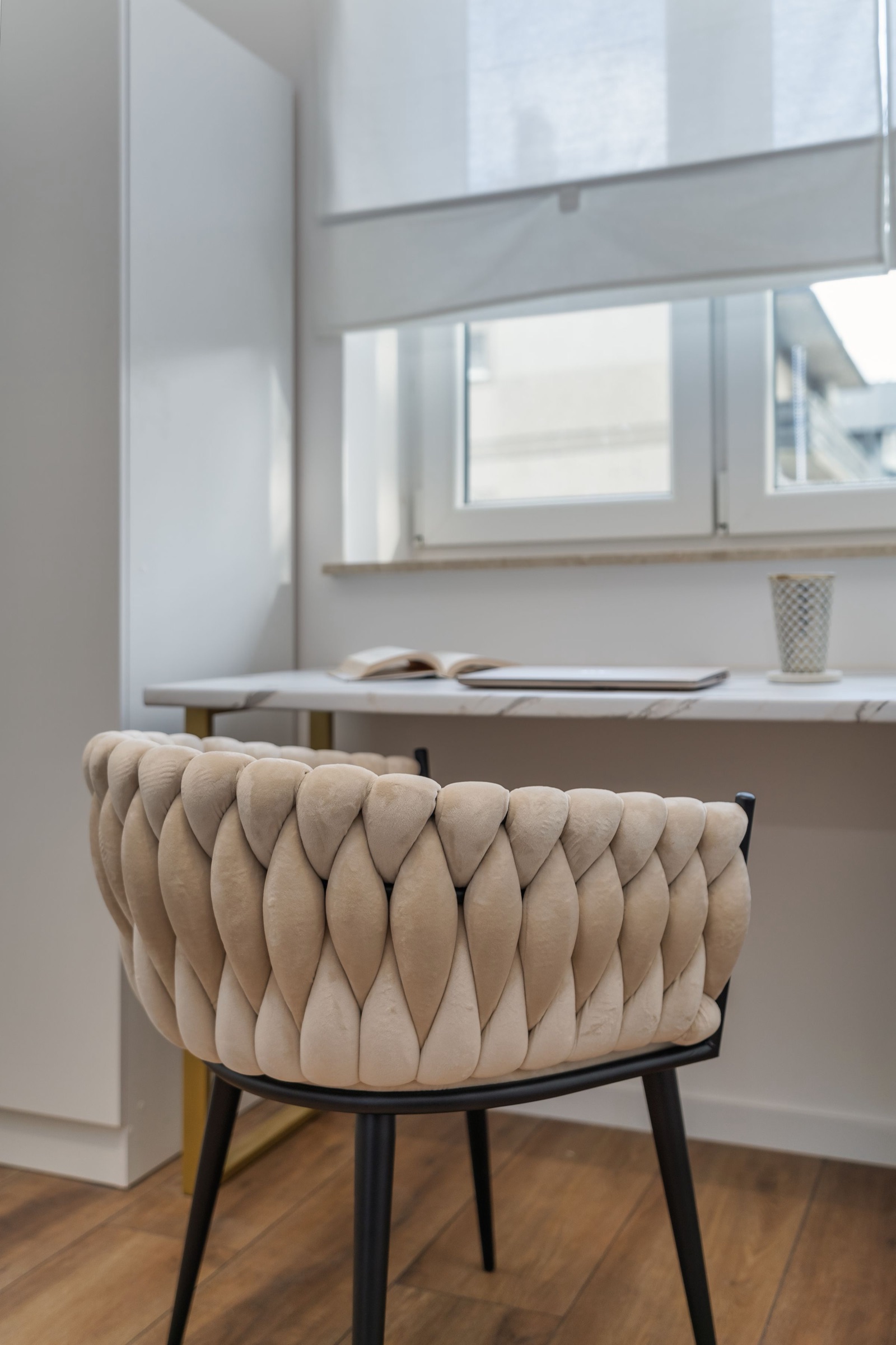 A marble-top desk on slim gold legs set under a window with a white fabric roller blind half down, an open book, a closed laptop and a mosaic cup on the desk, and a beige woven-velvet chair in the foreground on a wood-look floor.
