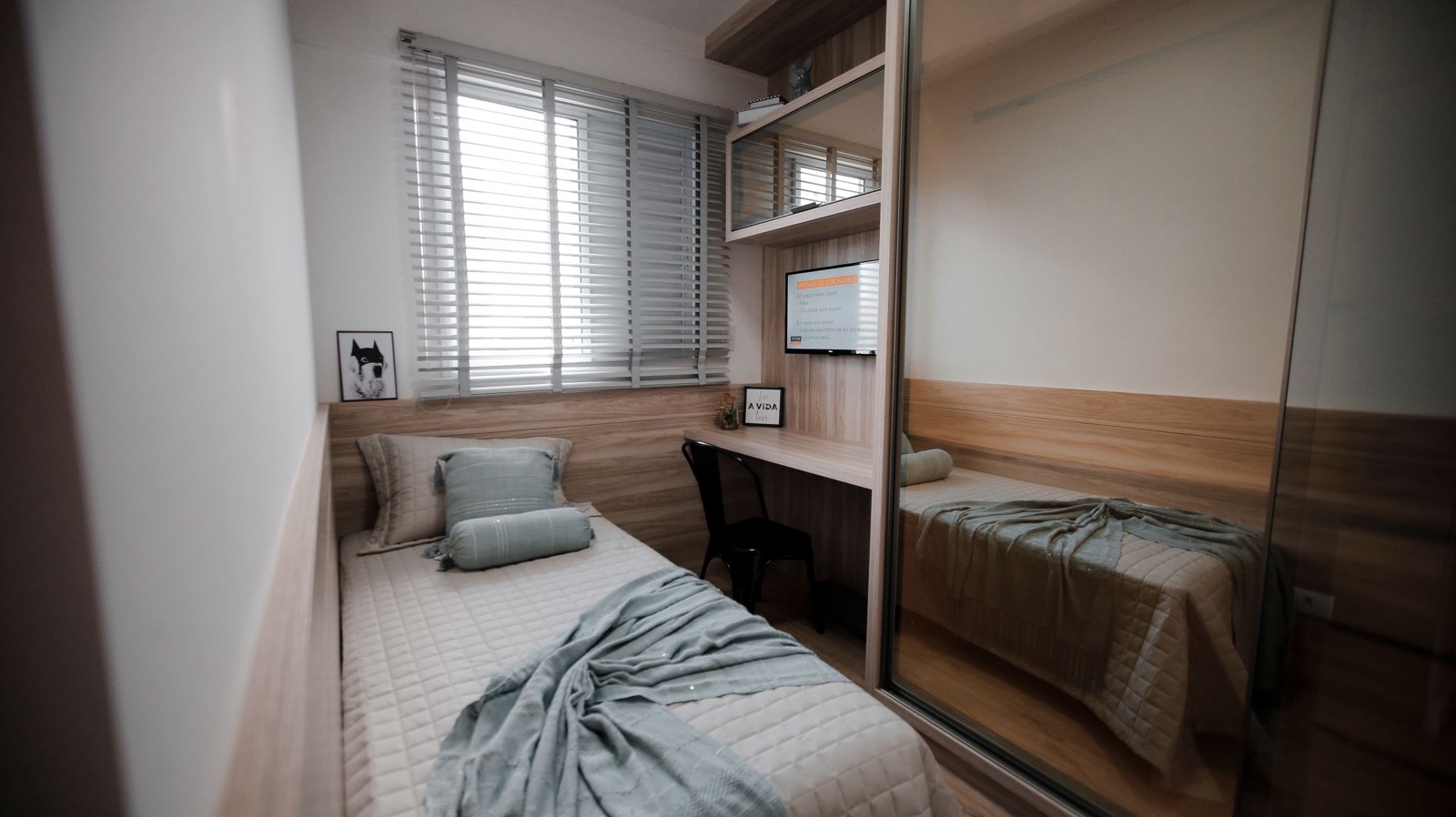 A narrow second bedroom with a single bed along one wall under a window with white blinds, a built-in wooden desk and chair at the window wall, a wall-mounted screen and shelves above the desk, and a full-height mirrored sliding wardrobe on the right reflecting the bed