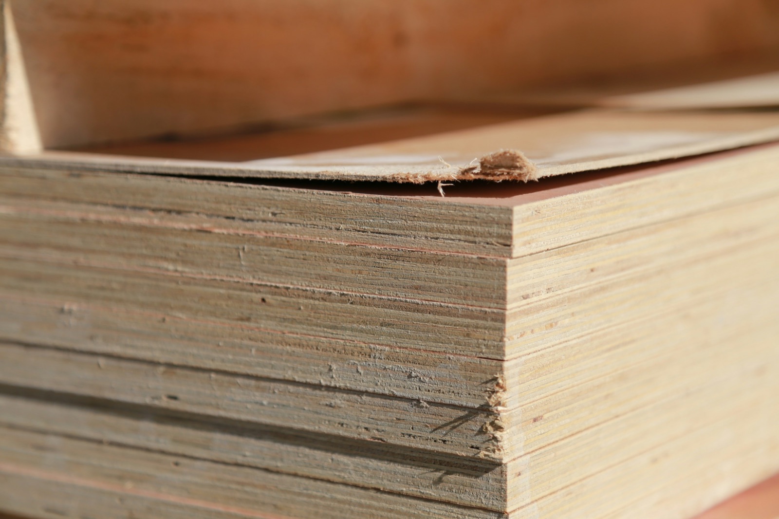 Close-up of the corner of a stack of plywood sheets, showing the thin glued plies on each cut edge and a reddish face veneer, with one top veneer torn and lifting at the corner