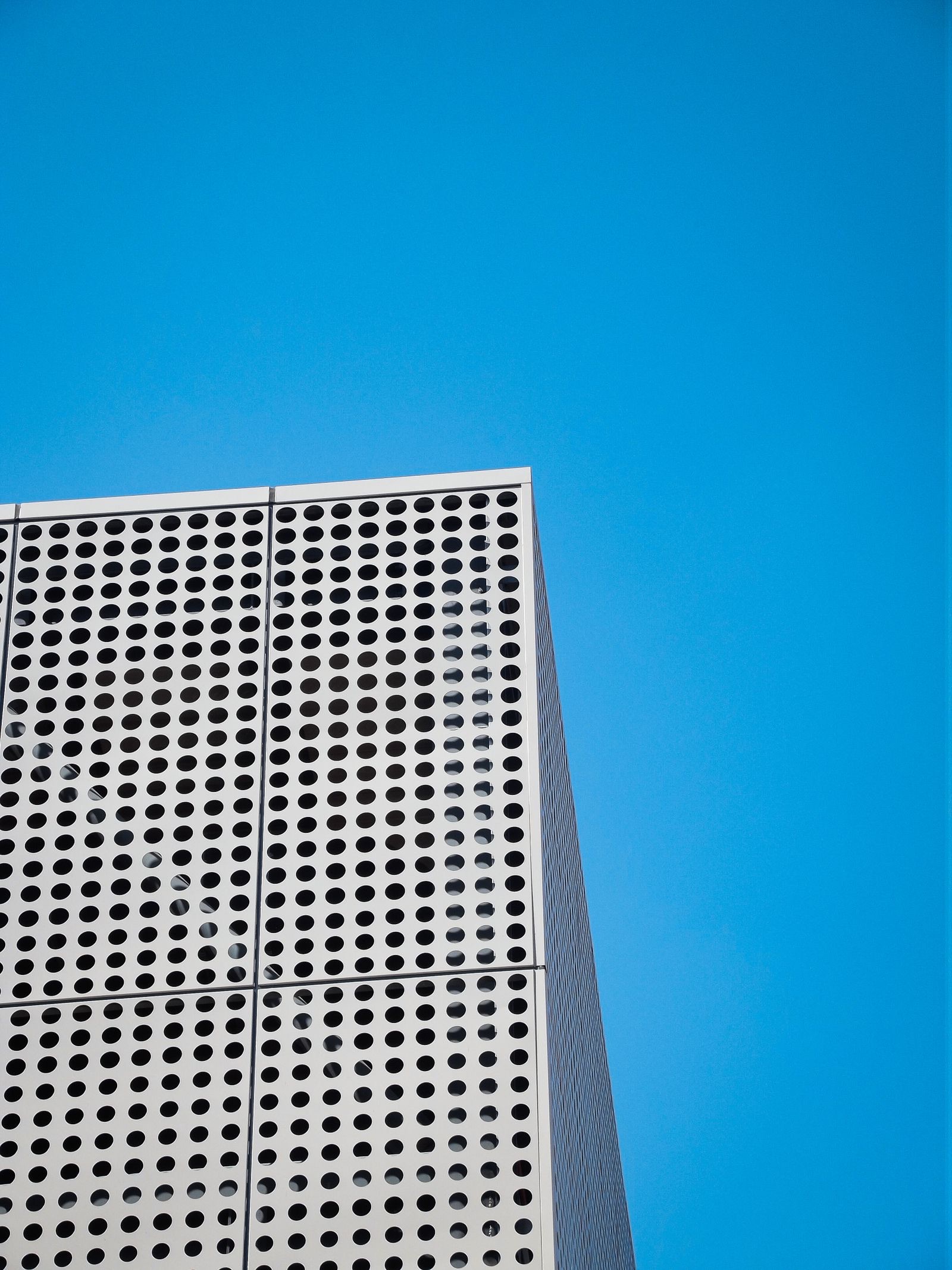 Corner of a building clad in pale grey perforated metal panels with rows of round holes, set against a clear blue sky.