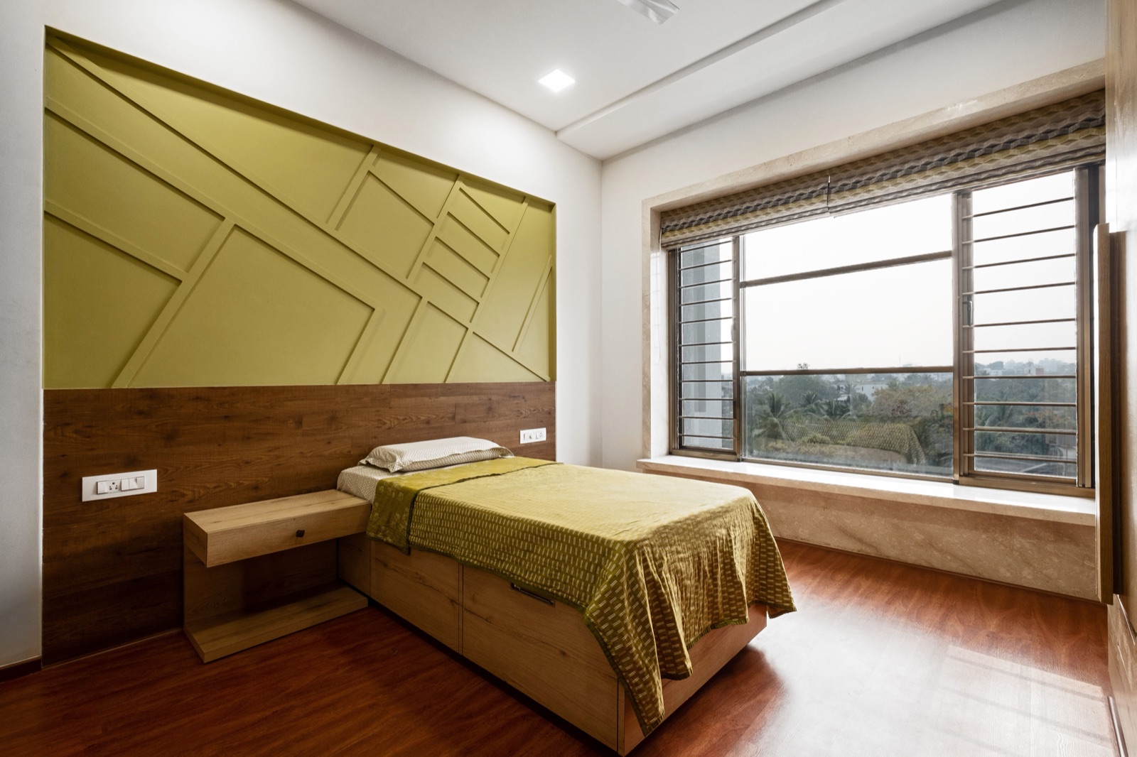 A bedroom in Pune with a single bed on a wooden base with pull-out drawers, a floating bedside drawer, an olive-green panelled wall above a wood-finish headboard band, and a wide window with a deep stone seat looking over trees