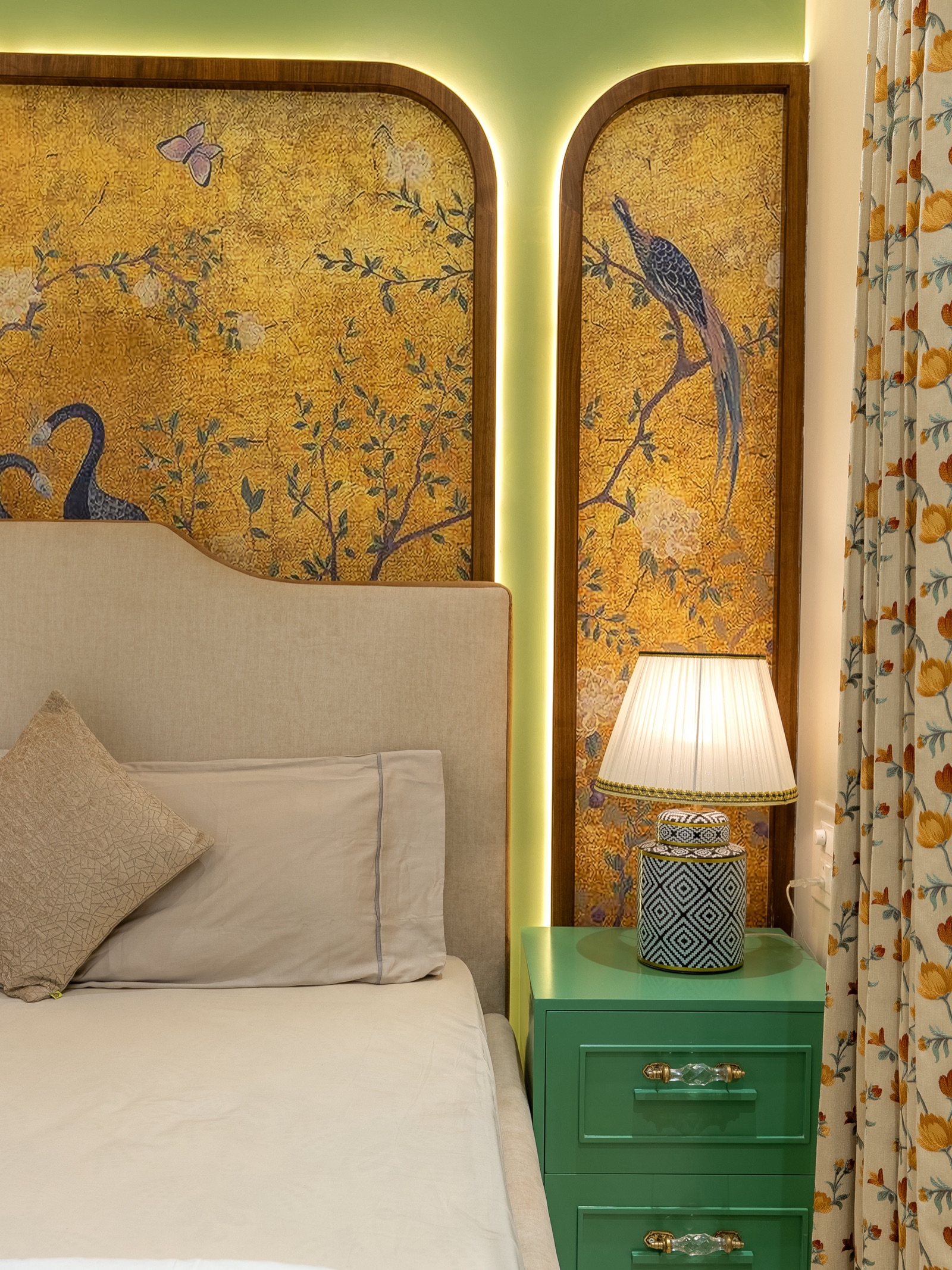 Photograph of a bed corner with gold botanical panels showing peacocks, set in round-topped wooden frames with warm LED backlighting on a pale green wall, a beige upholstered headboard, a green bedside chest and a pleated table lamp.