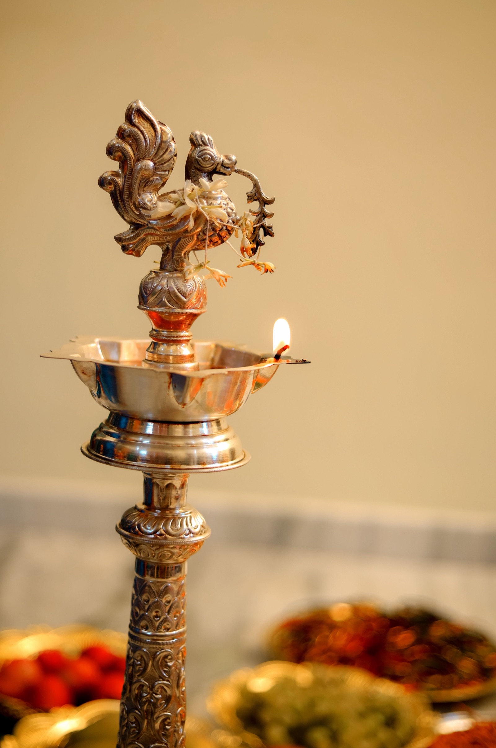 A tall silver-toned standing oil lamp with a peacock finial garlanded with jasmine, one wick lit, against a plain cream wall with brass plates blurred in the foreground.
