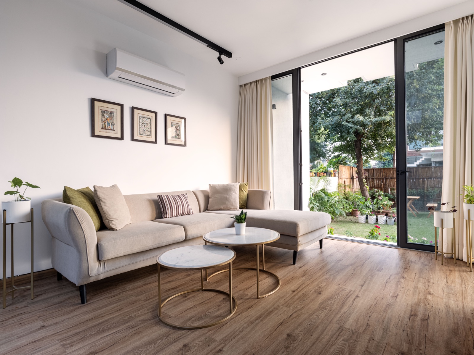 A bright living room with a beige L-shaped sofa, two round marble-topped nesting tables and a wood-toned plank floor running to full-height glass doors that open onto a garden.