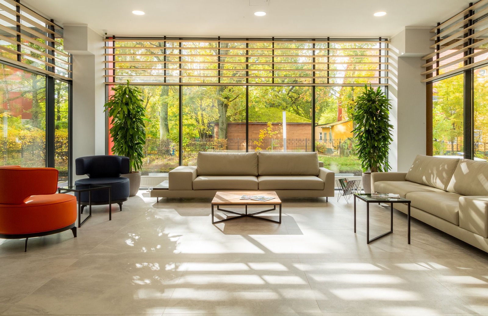 A bright lounge with a floor of large pale grey stone-look tiles in a wide grid, sunlight casting window shadows across it, beige sofas, an orange and a navy armchair, a timber coffee table and full-height glazing onto trees