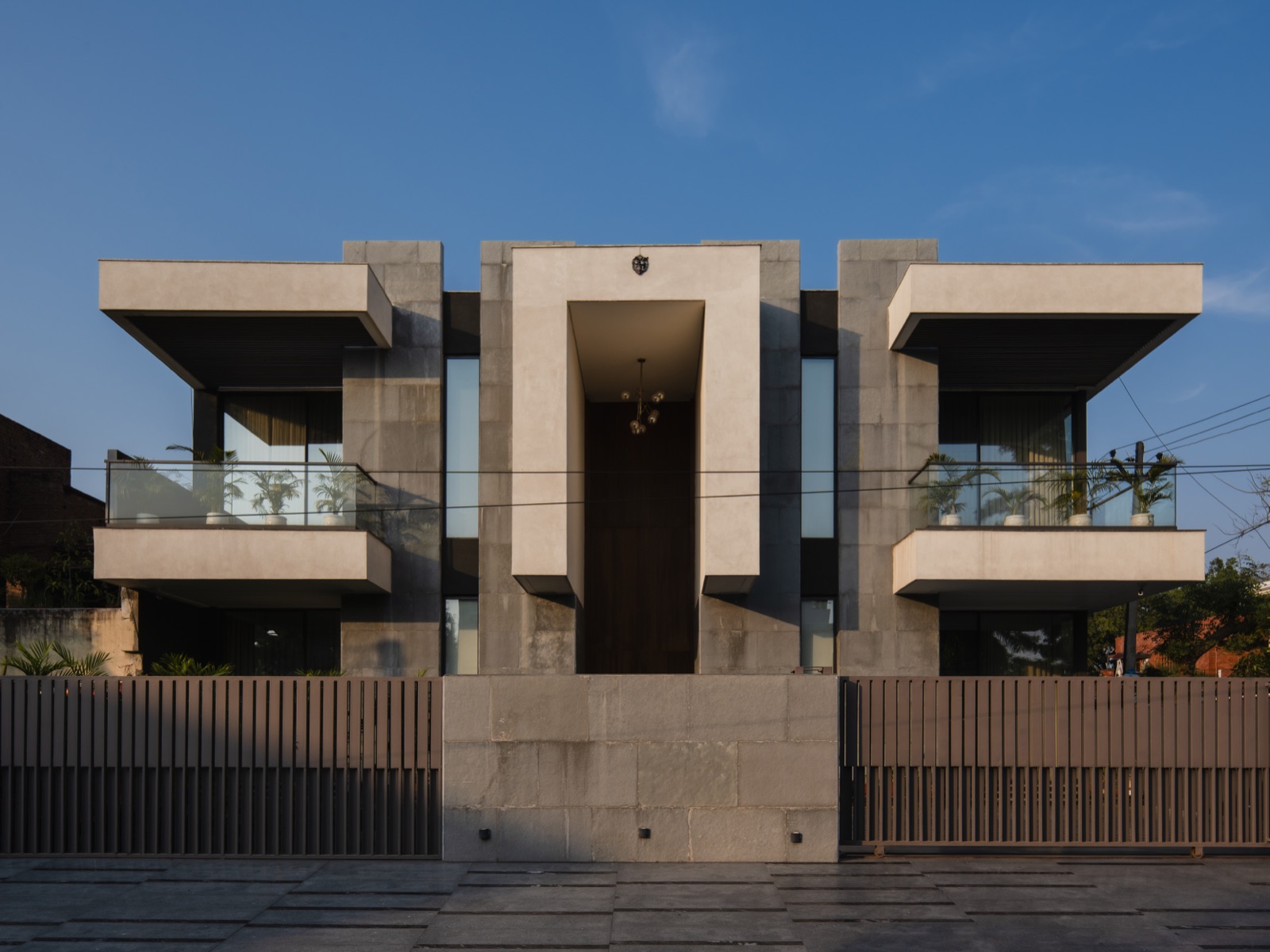 Symmetrical two-storey house in Panchkula seen straight from the street: grey stone-clad walls, a tall recessed entrance portal, two deep cantilevered balconies with glass railings, and a stone wall with slatted metal gates in front