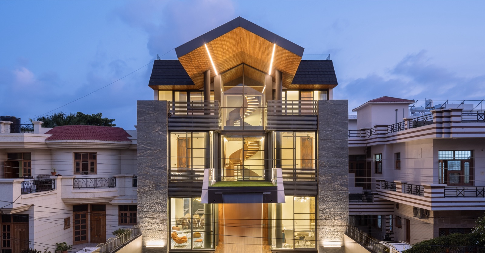 Narrow three-storey house in Panchkula at dusk, lit from within: a pitched roof with a timber soffit and two strip lights, full-height glazing showing a spiral stair, dark horizontal louvres at the balconies, and grey veined stone side walls washed by uplights, between two older neighbouring houses