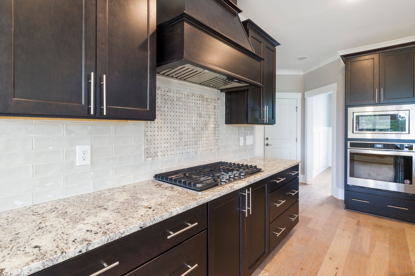 Kitchen with dark wood cabinets and a granite counter holding a five-burner gas hob. Pale glossy rectangular tiles cover the wall between counter and cabinets, and a framed panel of small basket-weave mosaic fills the wall behind the hob up to the wooden chimney hood.