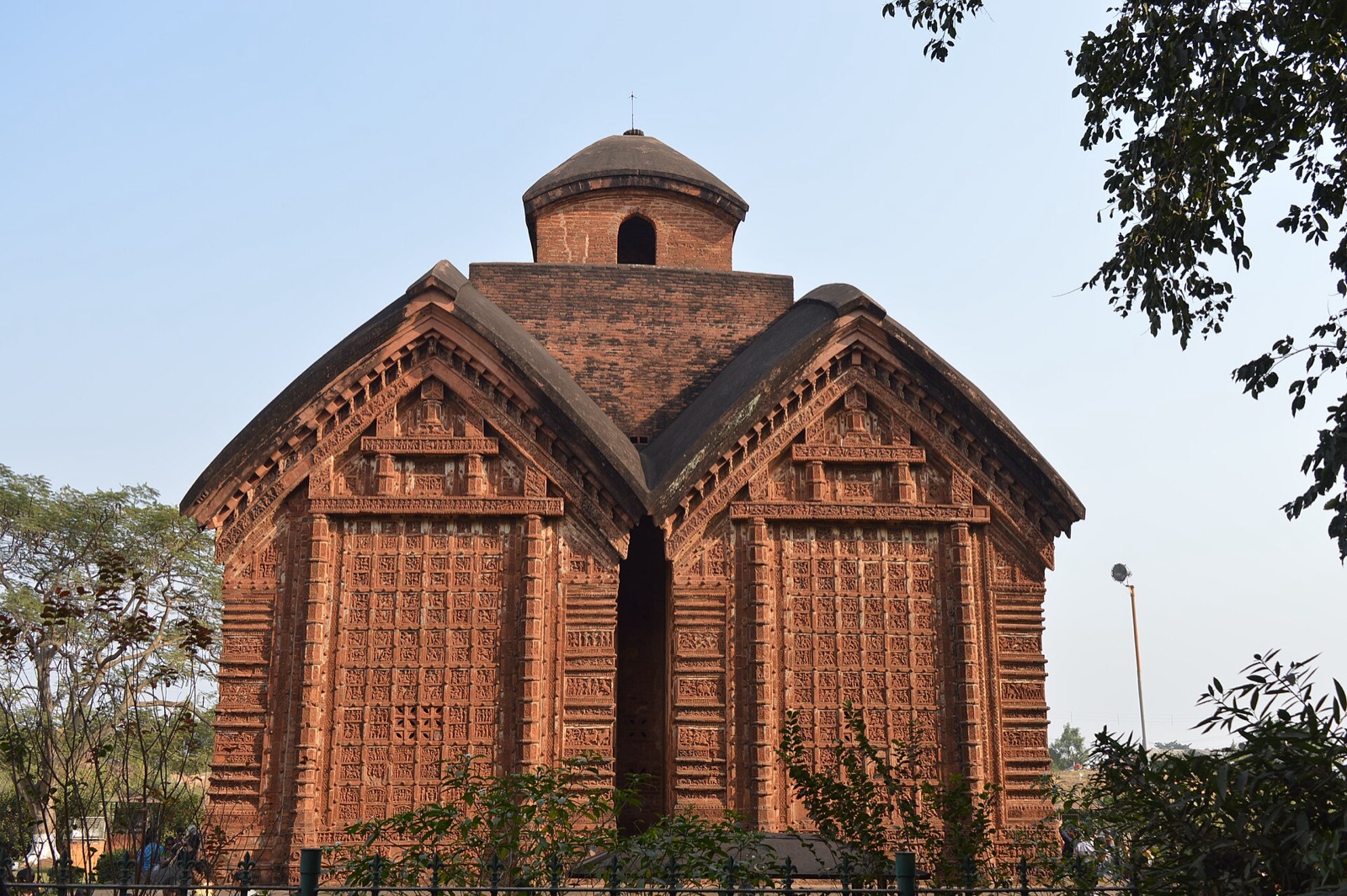 A brick temple seen from the front: two curved, hut-like roofs joined side by side, crowned by a small domed tower, with every wall face covered in a grid of carved red terracotta panels, under a clear blue sky with trees on either side.
