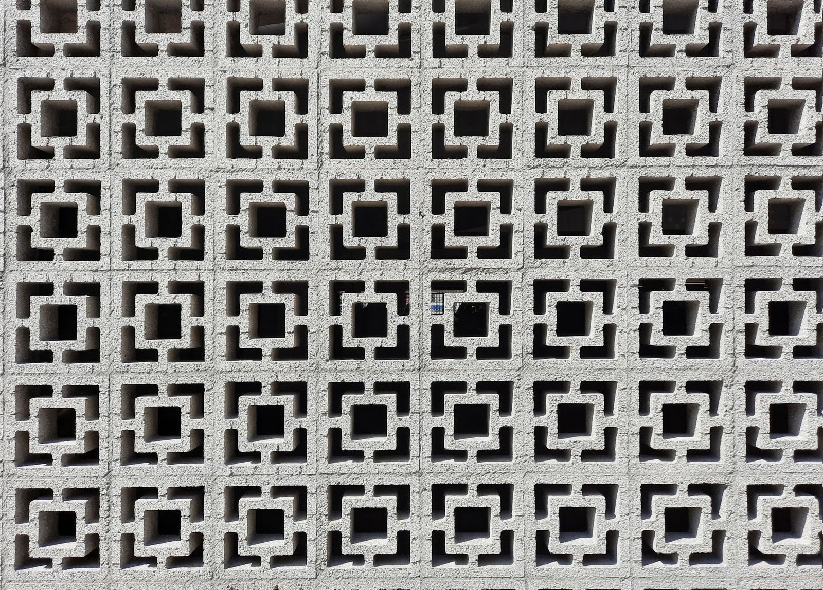 A wall of grey precast concrete breeze blocks, each square block with a square opening framed by a stepped square border, laid in a straight grid so dark openings repeat across the whole wall.