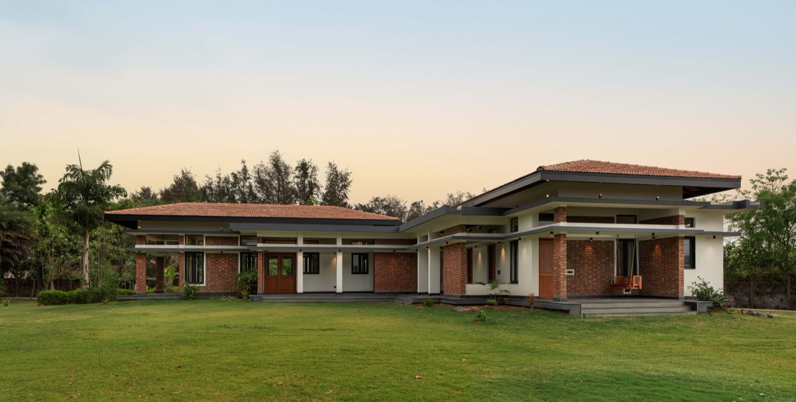 Single-storey farmhouse with exposed red brick walls and piers, white plastered bands, deep grey overhangs and clay-tiled pitched roofs, set on a wide lawn at dusk