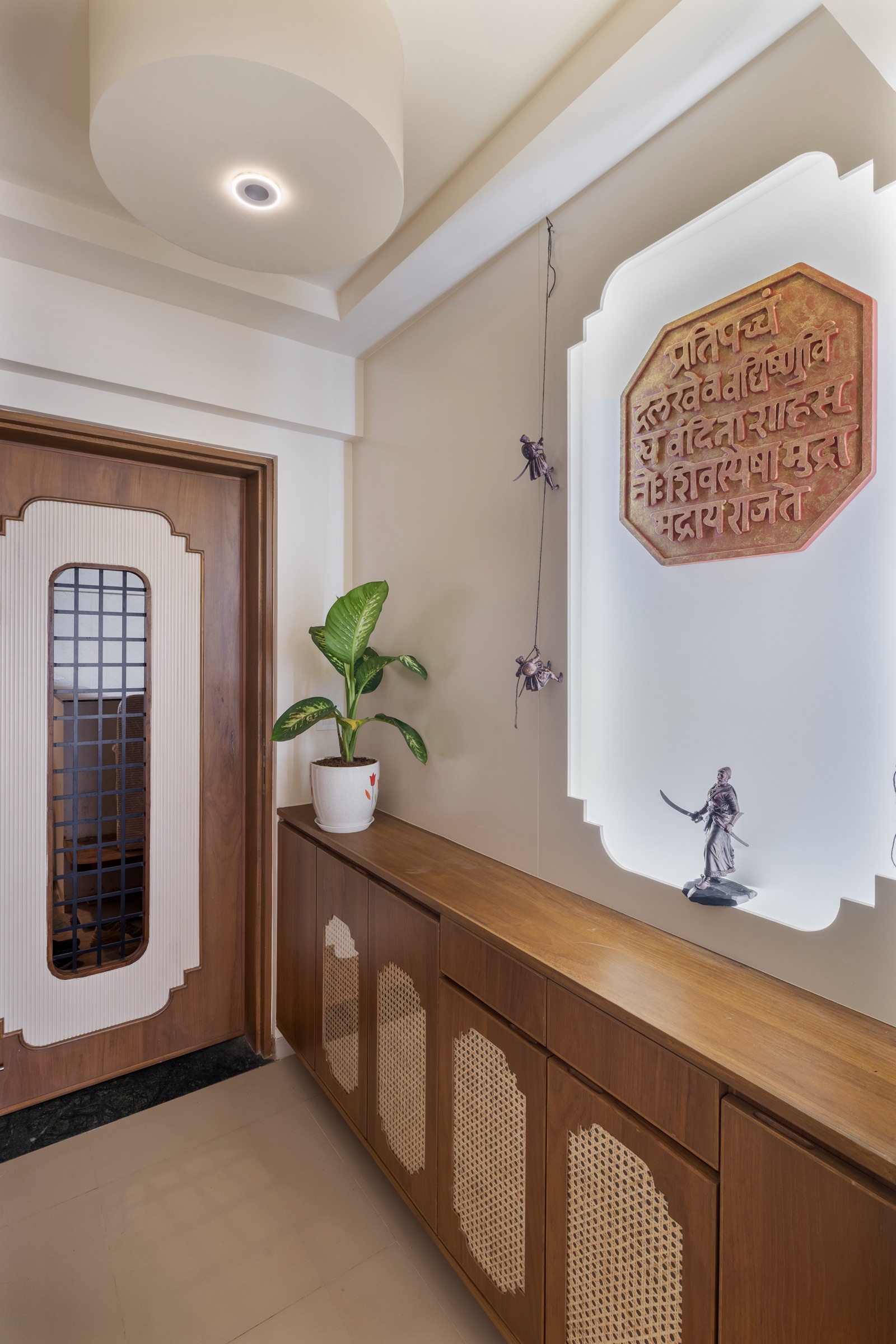 Home entrance with a wood-veneer main door that has a white fluted inset panel and a narrow black metal grill vision panel, beside a long wooden shoe cabinet with cane-backed shutters and a backlit niche