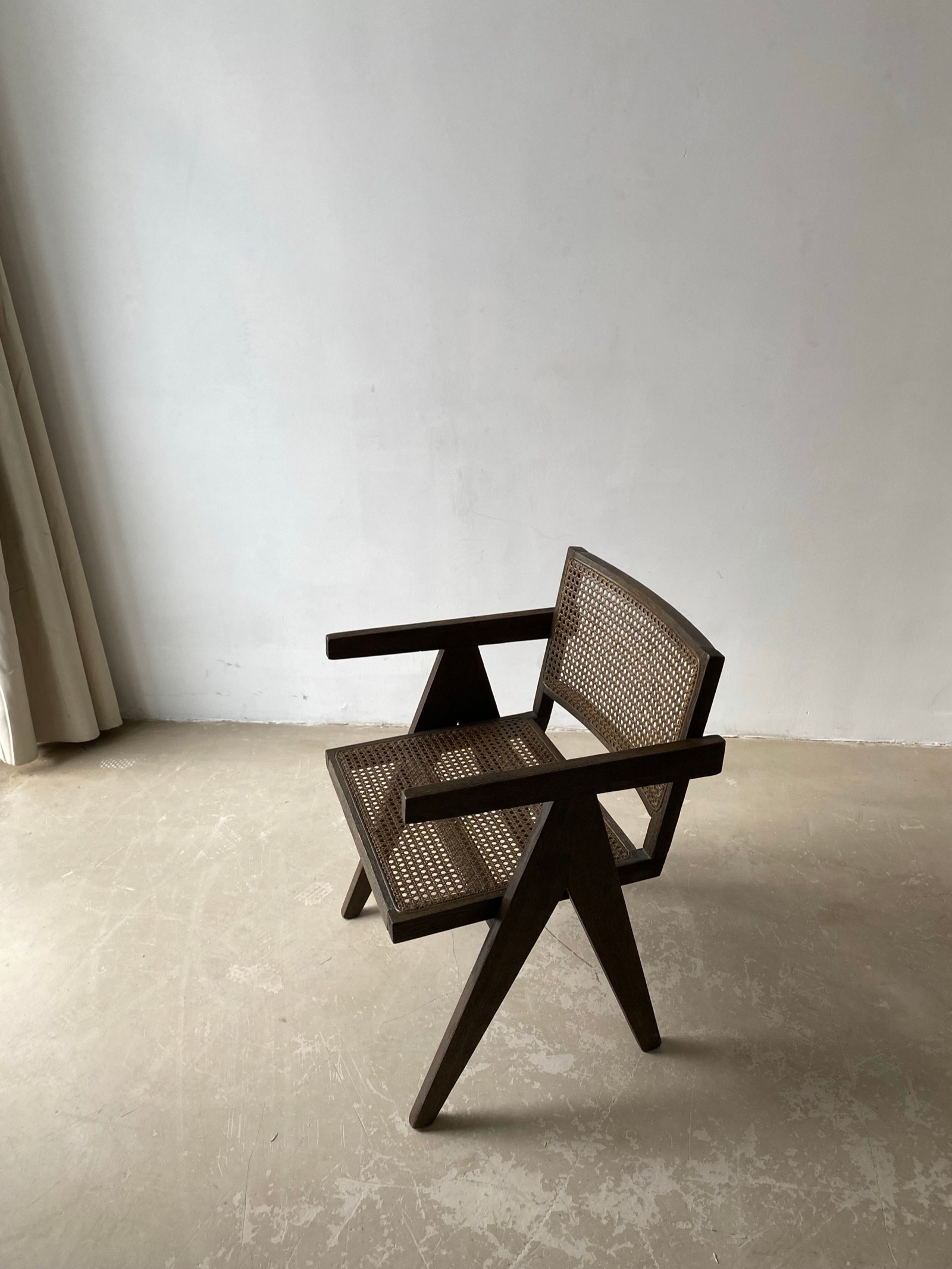 Dark-stained wooden armchair with a woven cane seat and back, straight arms and V-shaped legs, standing on a bare concrete floor beside a white wall and a cream curtain