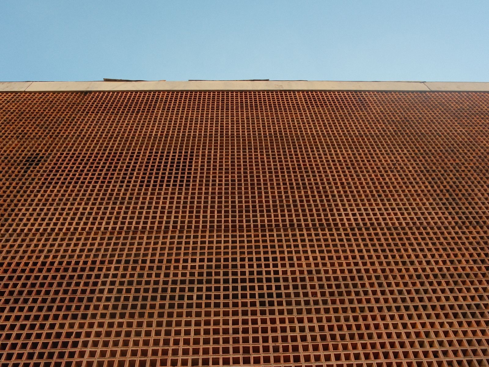 Looking up at a tall facade made entirely of small square terracotta openings in a fine even grid, with a pale concrete coping along the top and a clear blue sky above.