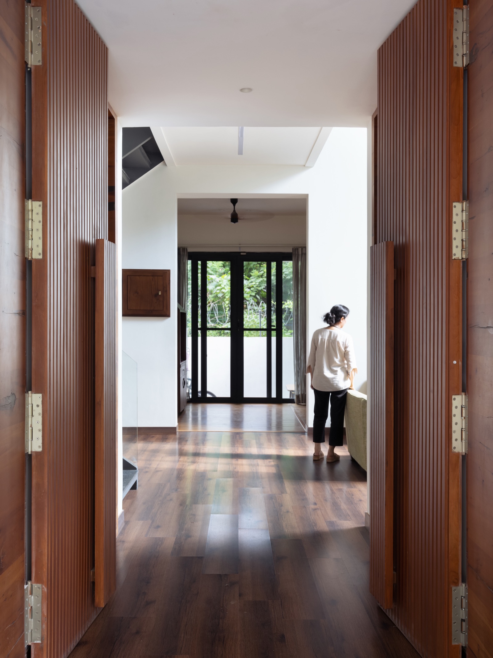 View through a pair of open fluted wooden doors with long timber pull handles and brass hinges, into a hallway with dark wood flooring leading to a bright living room