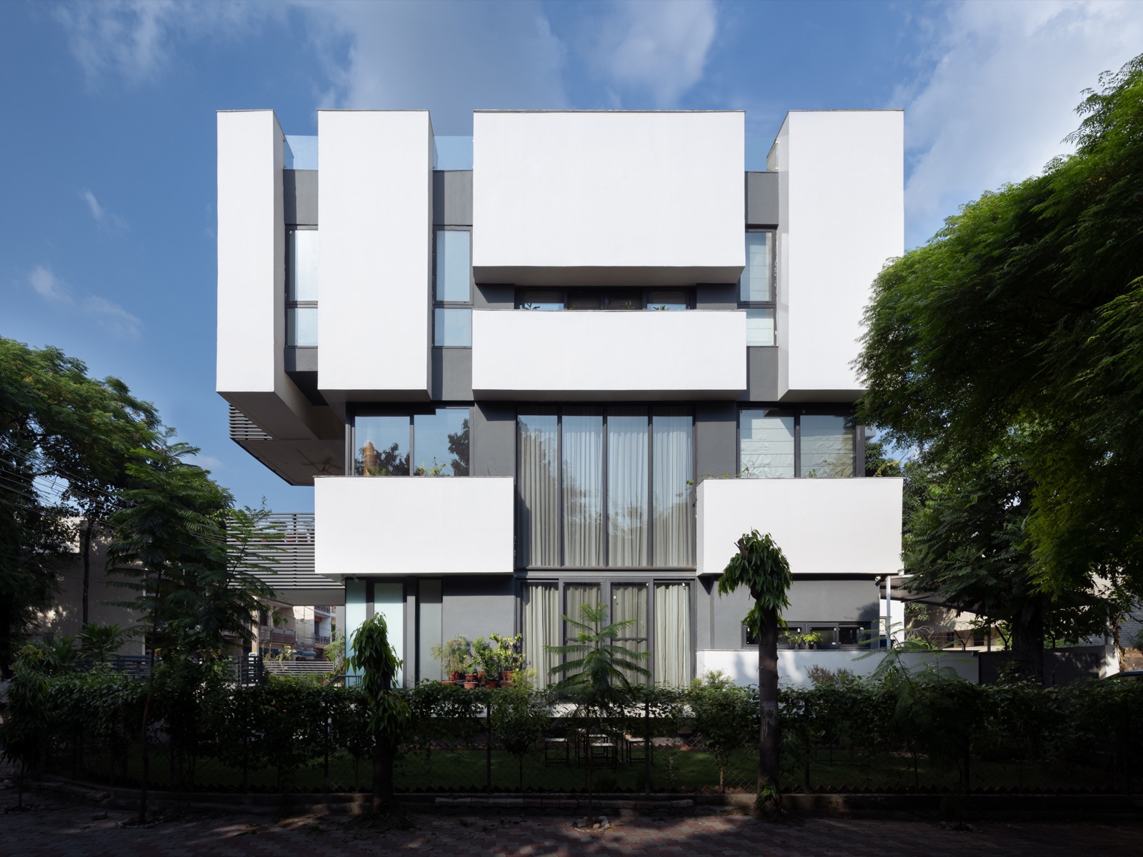 Street facade of Lacuna House in Panchkula, a white modern home with projecting white volumes, tall glazing and recessed terraces behind a hedge