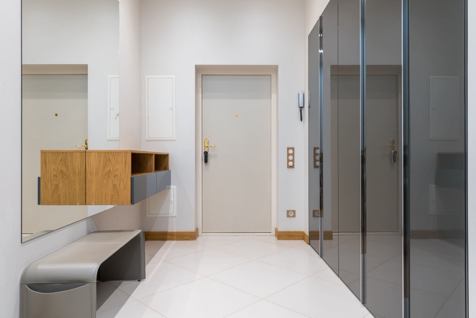 A white entrance hall with a wall-hung oak cabinet with two doors, two open cubbies and grey drawers fixed to a full-length mirror, a grey leather bench below it, a cream main door and a row of tall dark glass wardrobe doors on the right