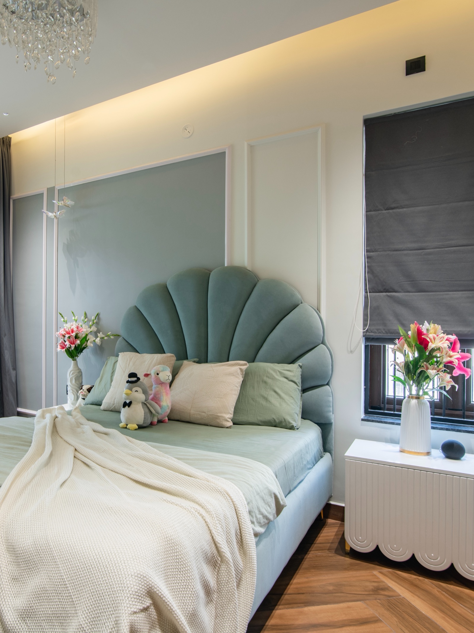 Girl's bedroom with pale grey-green panels set inside white wall moulding, a teal velvet scalloped headboard, sage bedding, a white fluted bedside table and a grey roman blind