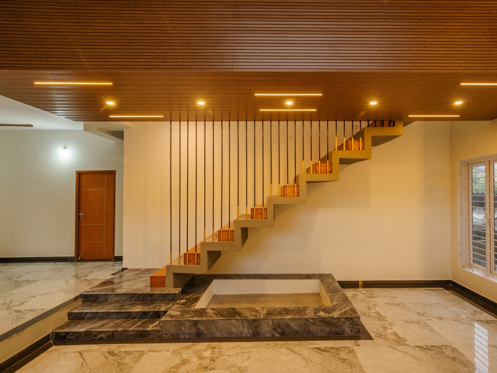 A straight staircase with timber treads on a folded concrete stringer rising along a white wall, with slim vertical rods dropping from a slatted wooden ceiling and a stepped grey marble platform at its foot.