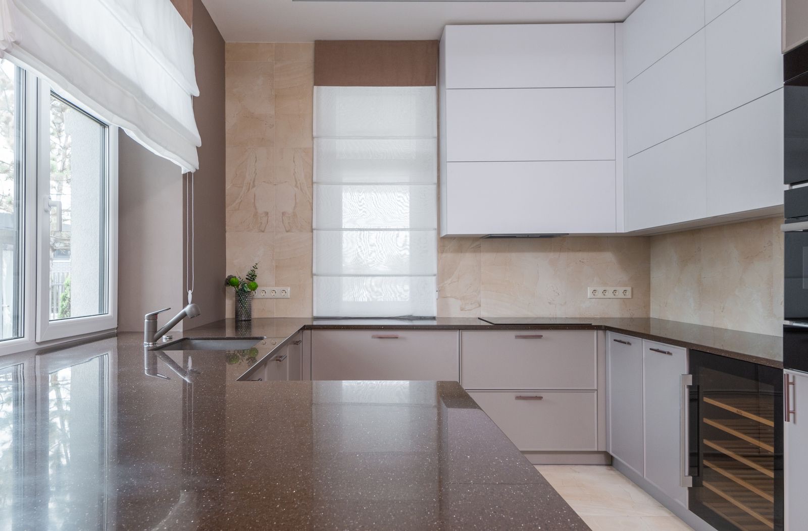 U-shaped kitchen with a polished brown speckled engineered-stone platform, an undermount stainless steel sink under a large window, light grey handleless drawers and a beige stone backsplash running up to white wall cabinets.