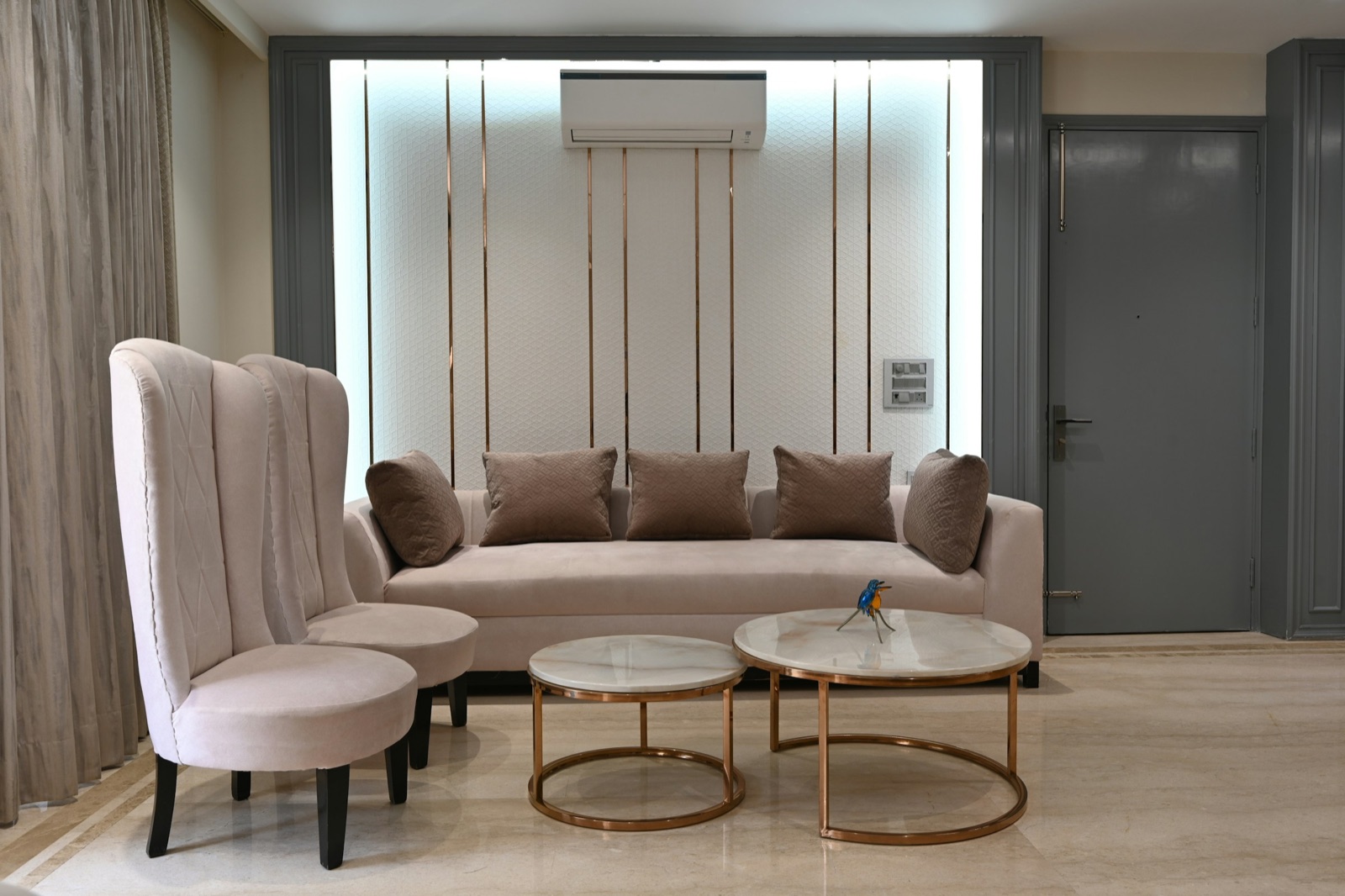 A pale pink three-seater sofa with brown cushions against a backlit panelled wall with copper strips and a split AC above, two tall high-backed armchairs on the left, and a pair of round marble-topped nesting tables with copper bases in front. A grey main door with a long handle is on the right and a marble floor runs throughout.