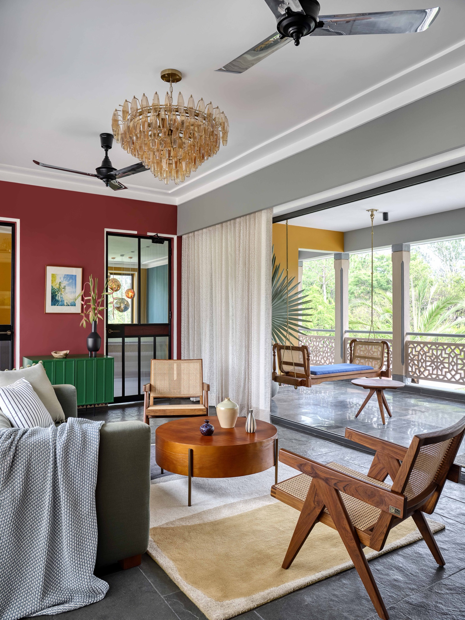 A living room with a deep red accent wall, a green sideboard, a grey sofa and cane armchairs around a round wooden table on a pale rug, slate floor, and a verandah beyond with an ochre yellow wall and a wooden swing
