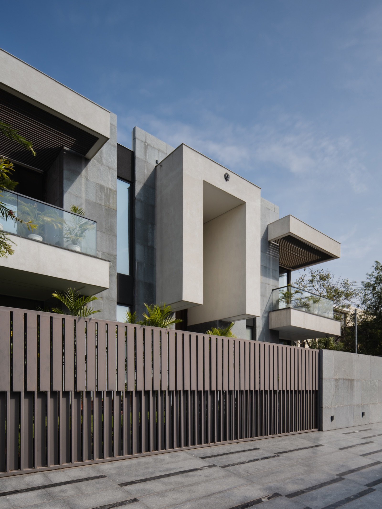 Street front of a contemporary two-storey house whose walls and boundary wall are clad in large grey stone slabs, with pale plastered cantilevered balconies, glass railings, a slatted metal gate and grey stone paving in front